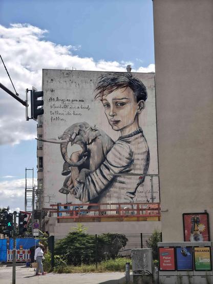 Photo of the side wall of a house.
Hera (the artist) drew a huge boy holding an elephant on the wall with the words: "As long as you are standing give a hand to those who have fallen" next to it.